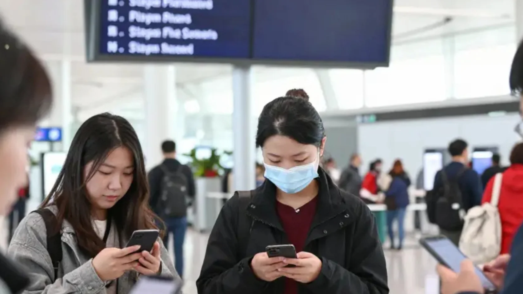 Passengers using smartphones for self-service flight rebooking during a disruption, with flight information displays in the background.